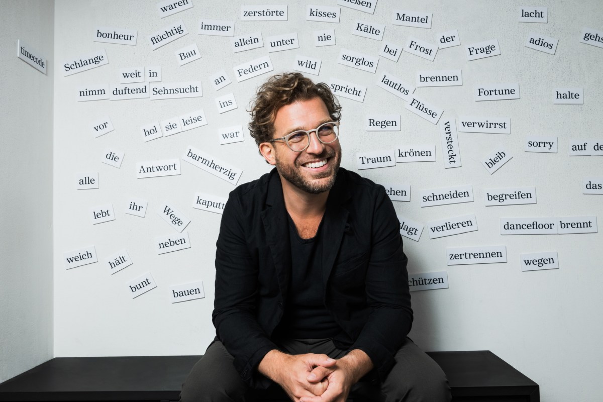 Julian Prégardien sitzt lächelnd vor einer Wand mit vielen aufgebrachten Wörtern wie "Sehnsucht", "begreifen", "bunt",