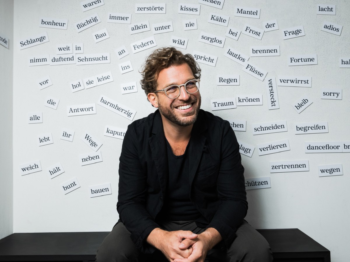 Julian Prégardien sitzt lächelnd vor einer Wand mit vielen aufgebrachten Wörtern wie "Sehnsucht", "begreifen", "bunt",
