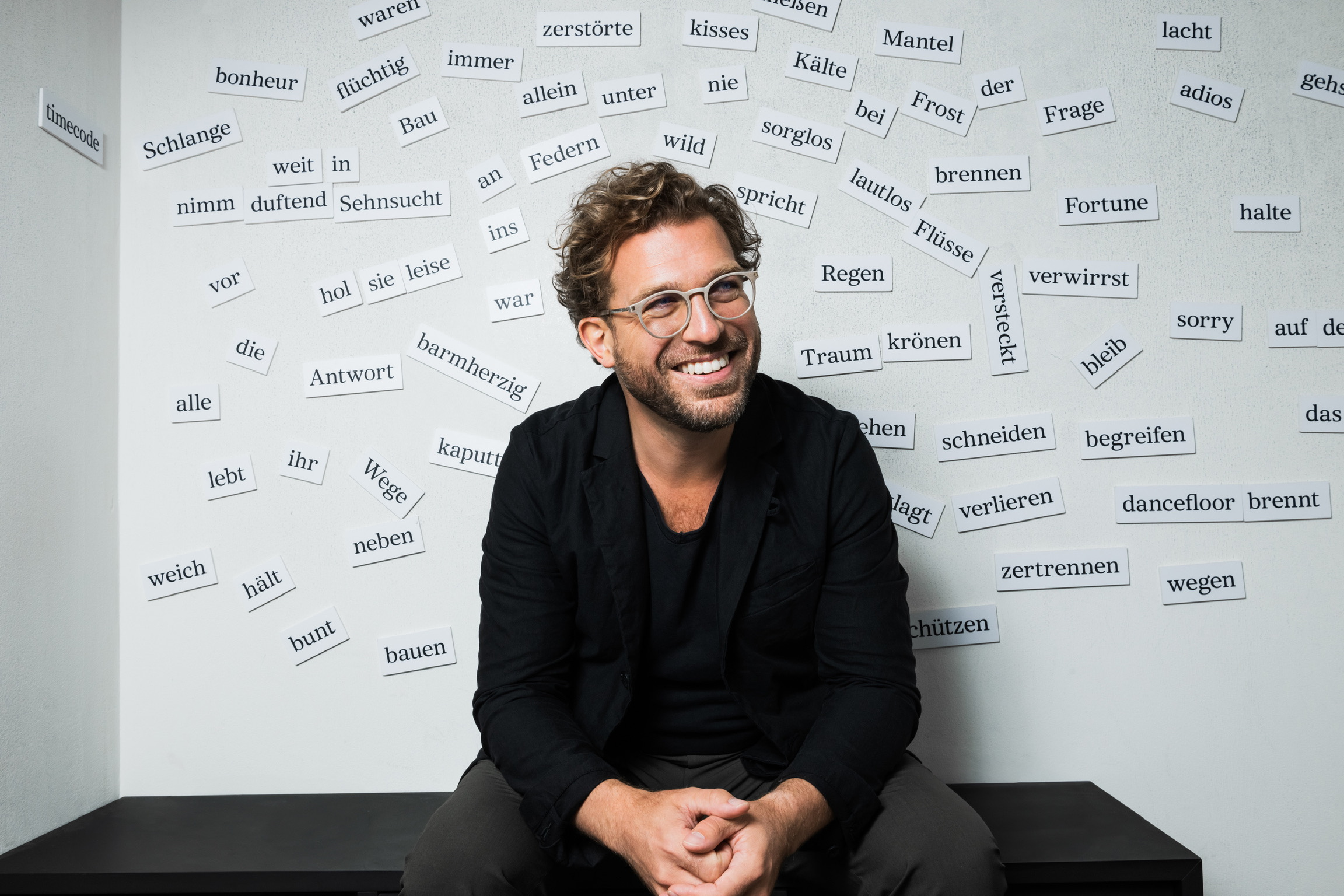Julian Prégardien sitzt lächelnd vor einer Wand mit vielen aufgebrachten Wörtern wie "Sehnsucht", "begreifen", "bunt",
